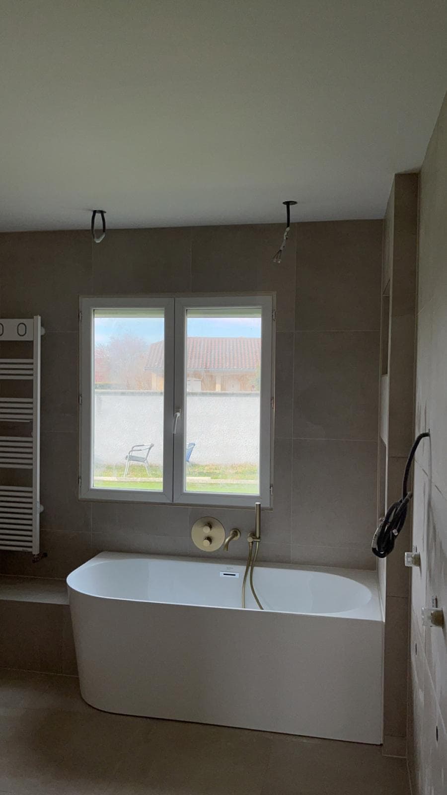 Baignoire adossée sous fenêtre à La Motte-Servolex, carrelage beige aspect pierre au sol et aux murs, robinetterie encastrée or brossé, luminaires pas encore posés