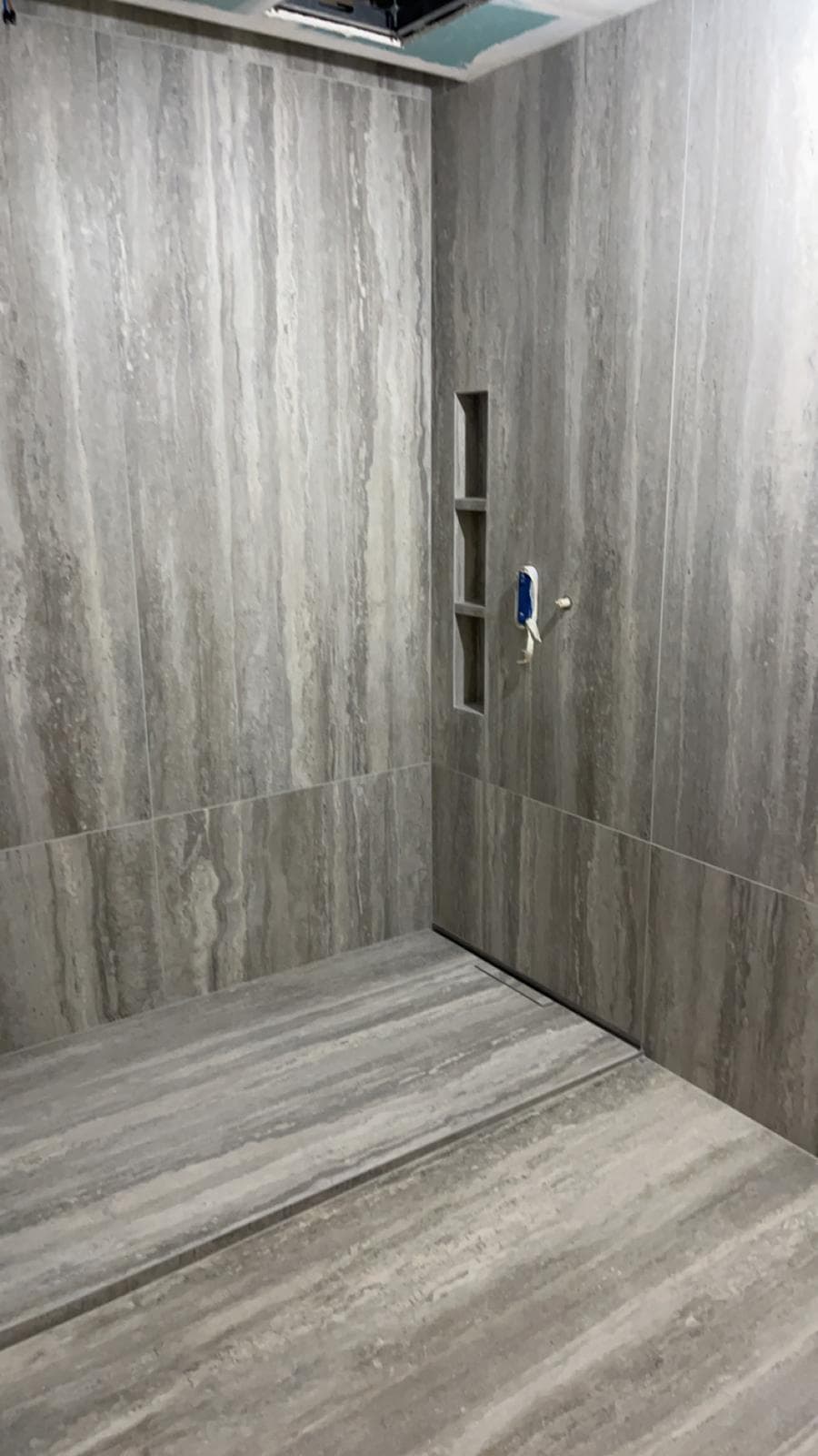 Douche à l’italienne en cours de finition à Gex : grès cérame très grand format effet travertin gris, caniveau linéaire et niche verticale à trois compartiments