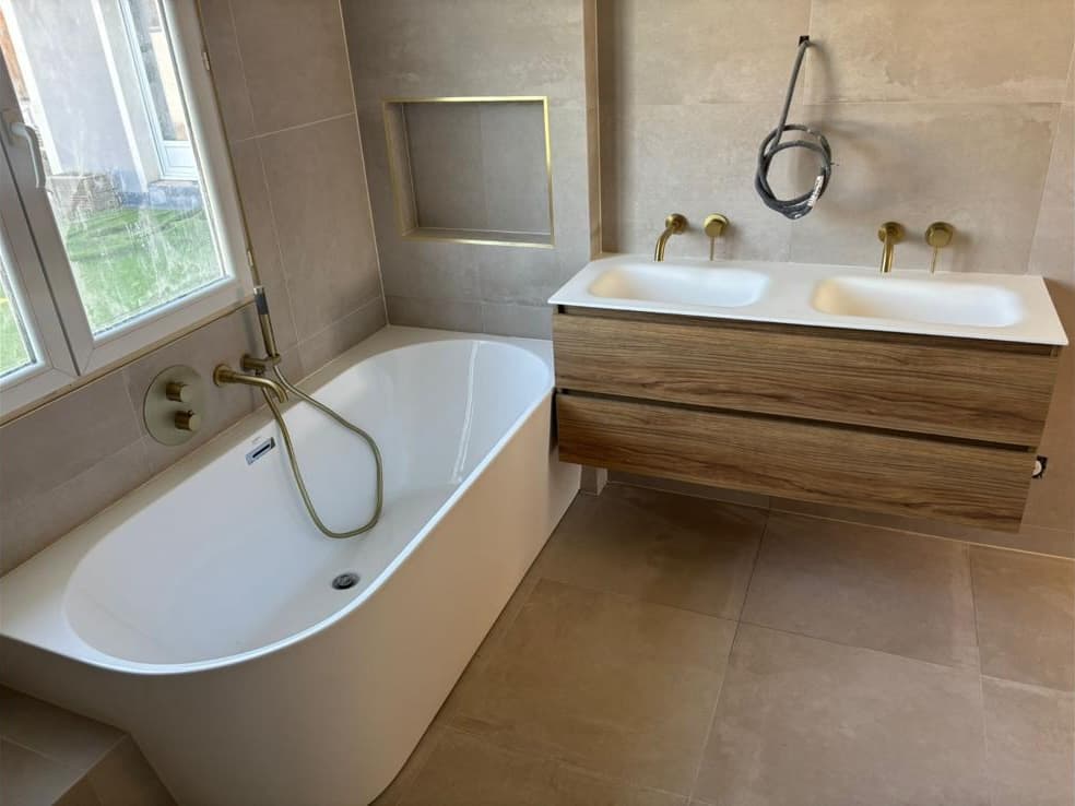Salle de bain terminée à La Motte-Servolex : carrelage beige grand format, baignoire adossée, niche à profilé doré et meuble double vasque effet bois