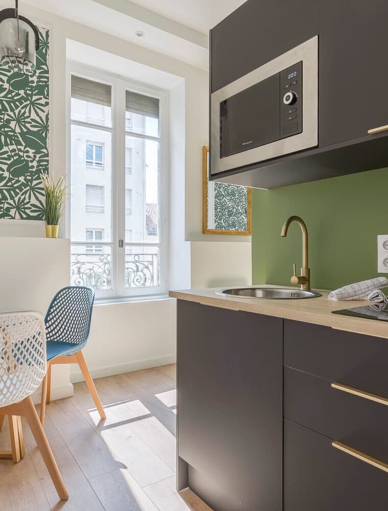 Kitchenette anthracite à robinet laiton et plan décor bois clair, mur peint vert olive, face à une porte-fenêtre à garde-corps en fer forgé, à Chambéry