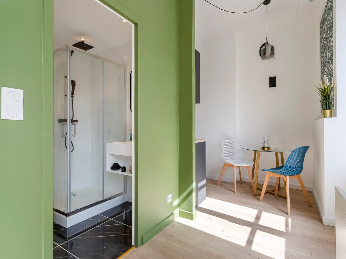 Salle d’eau d’un studio à Chambéry : cabine de douche vitrée à portes coulissantes sur receveur, sol en carrelage effet marbre noir veiné doré, cloison vert olive