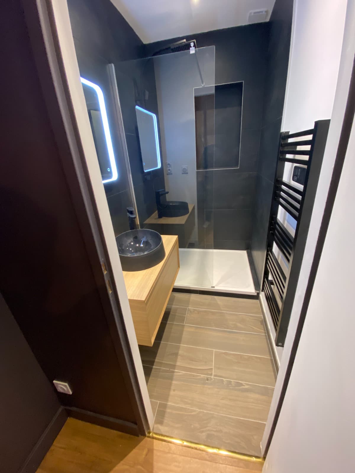 Salle de douche vue du couloir à Bourg-Saint-Maurice : sol en grès imitation bois, barre de seuil laiton, faïence anthracite et paroi de douche vitrée