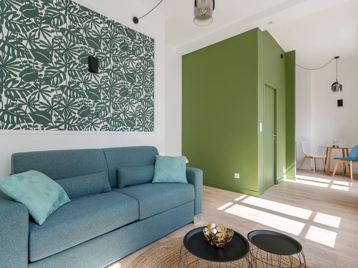 Séjour rénové à Chambéry : canapé bleu canard, panneau de papier peint tropical vert encadré de moulure et volume cloisonné peint vert olive avec porte intégrée
