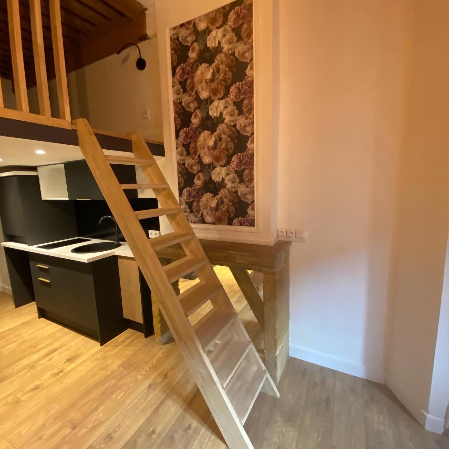 Échelle de meunier en bois vers une mezzanine sous poutres anciennes, kitchenette noire et cheminée en pierre dans un studio rénové à Annecy