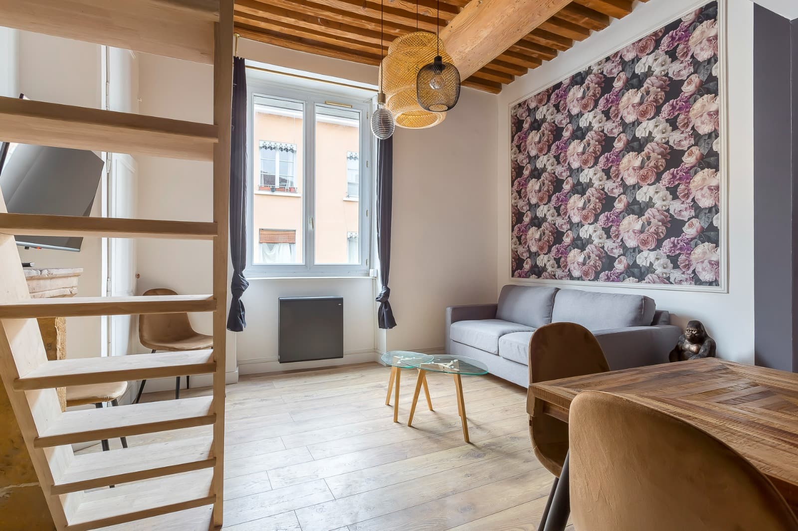 Séjour d’un appartement ancien rénové à Annecy : plafond à la française à solives apparentes, suspensions en rotin et panneau de papier peint floral encadré